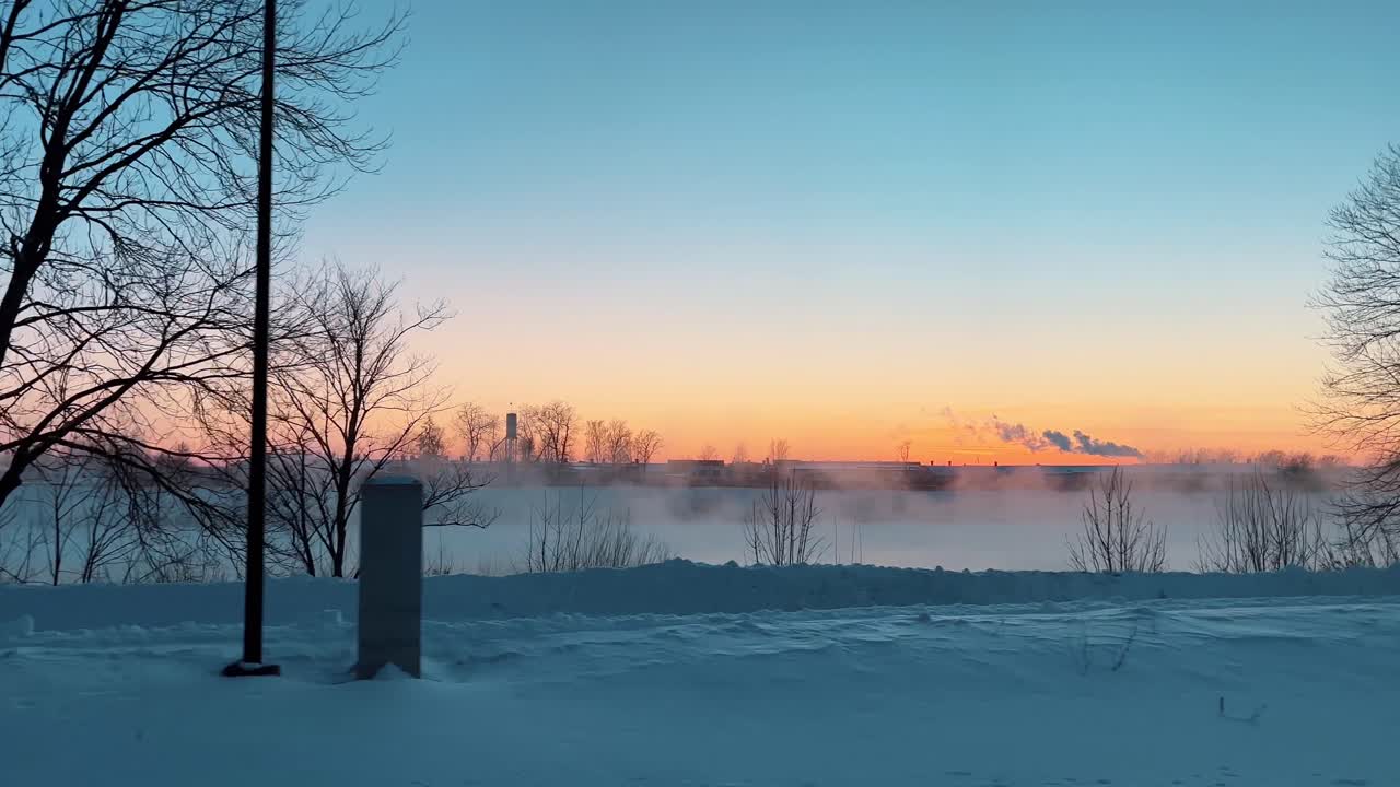 Passing by a serene city park covered in snow during a cold winter sunset, with thick fog rising from the river and bare trees silhouetted against the colorful morning sky