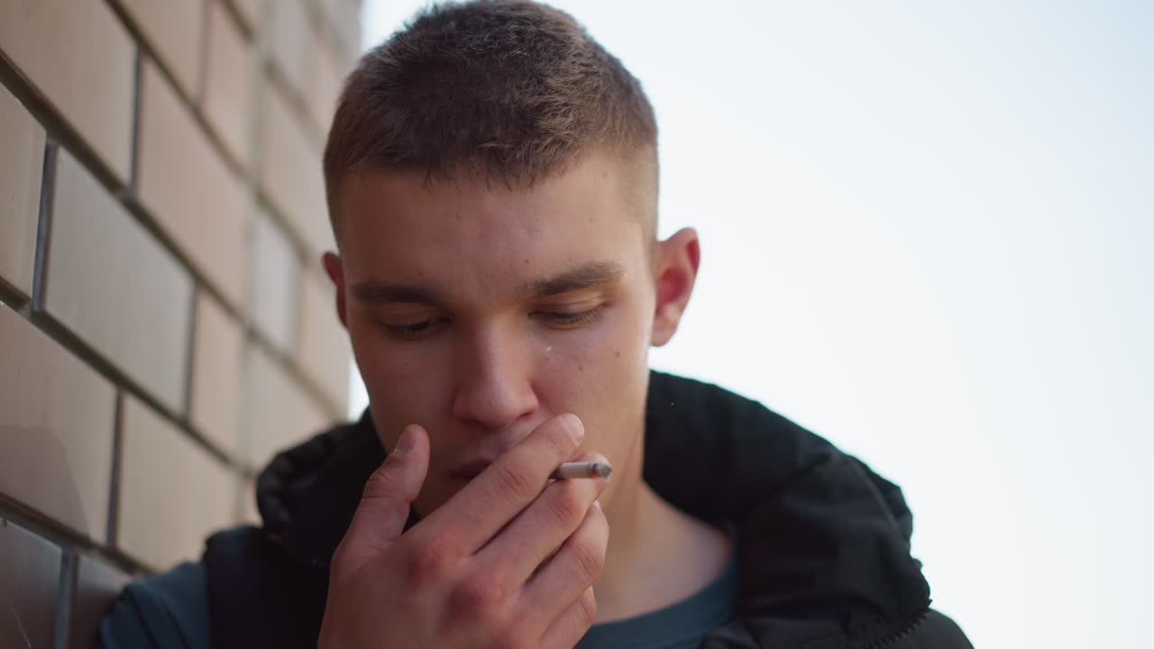 portrait view of fair skinned man smoking thoughtfully while leaning against brick wall, eyes closed in silent reflection with soft daylight casting shadow on textured surface behind him