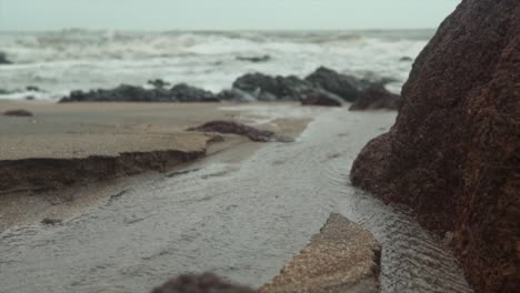 Agua-Que-Fluye-De-Regreso-Al-Océano-Entre-Piedras-En-La-Playa-Con-Olas-Rompiendo-En-La-Orilla-En-El-Fondo