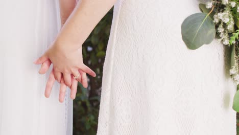 diverse female couple holding hands and bunch of flowers in garden