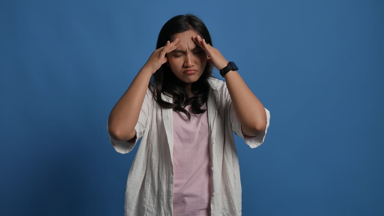 Stressed out young asian female suffering migraine headache, beautiful woman standing hand-over-head against isolated blue background.