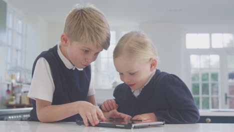 two children wearing school uniform in kitchen playing with digital tablet on counter