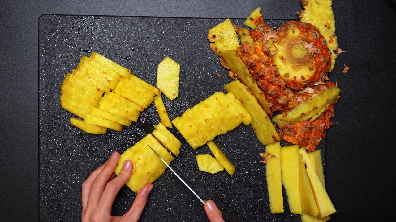 Vista dall'alto di ananas affettato su un tagliere scuro. Prospettiva dall'alto, polpa giallo vivido, buccia pelata, coltello in mano, pezzi sparsi, superficie umida.