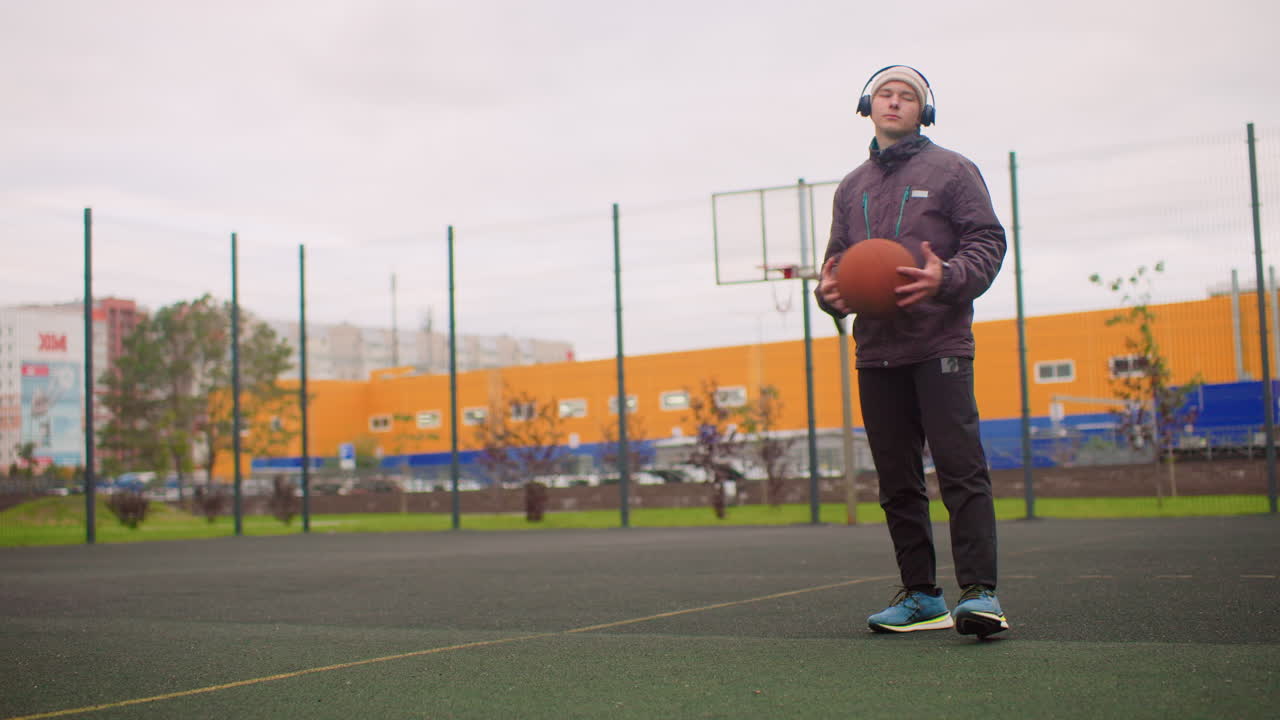 Caucasian man dribbling on outdoor court, wearing hoodie and headphones, bouncing basketball beside fenced hoop, orange warehouse backdrop, overcast sky, synthetic turf, focused expression, solo urban