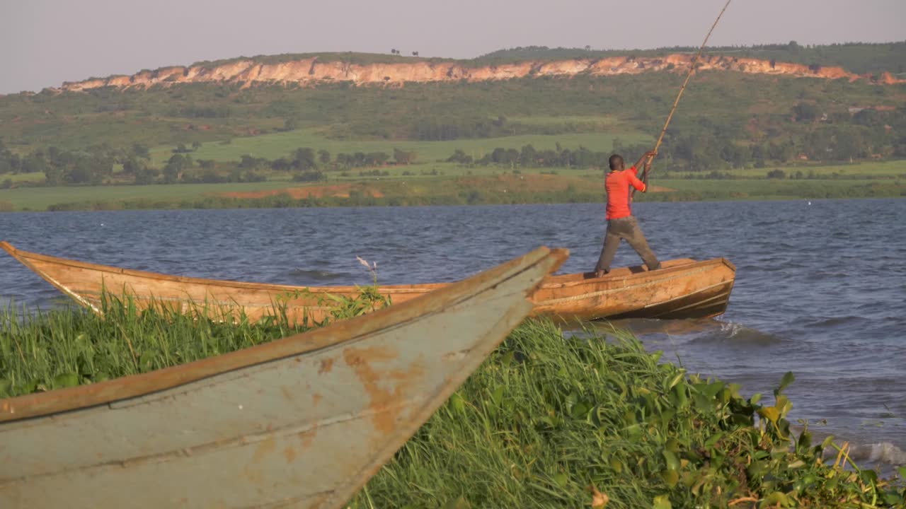 Slow motion shot of an African fishermen pushing his traditional wooden canoe with a large stick passed the shores of Lake Victoria.