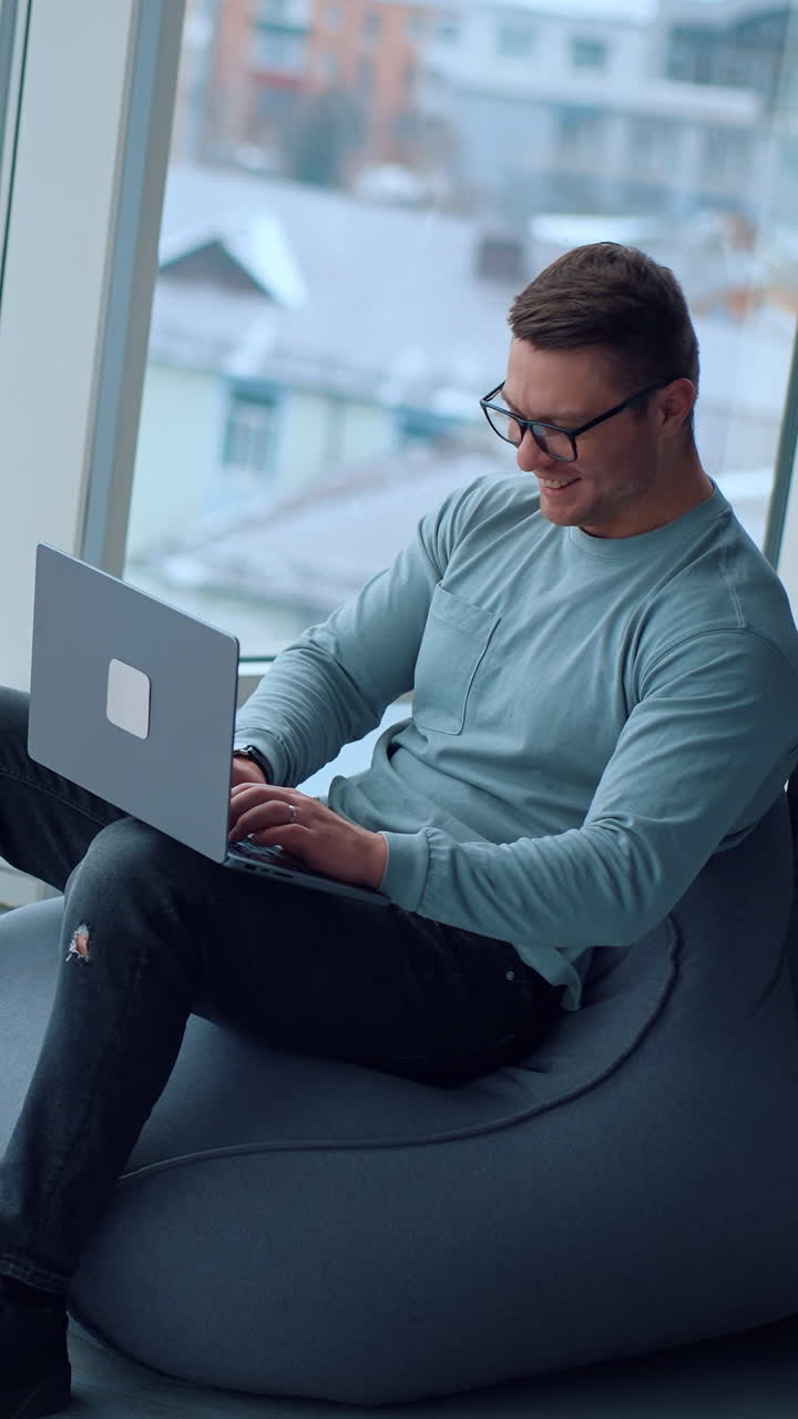 Comfortable work conditions in the modern office. Happy young man sitting in bean bag chair typing on his laptop at looking at window. High angle view. Vertical video