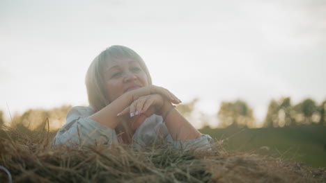 woman lying on piled hay with warm reflective smile, enjoying peaceful rural atmosphere, soft golden sunlight highlights her face, creating a calm and serene countryside moment