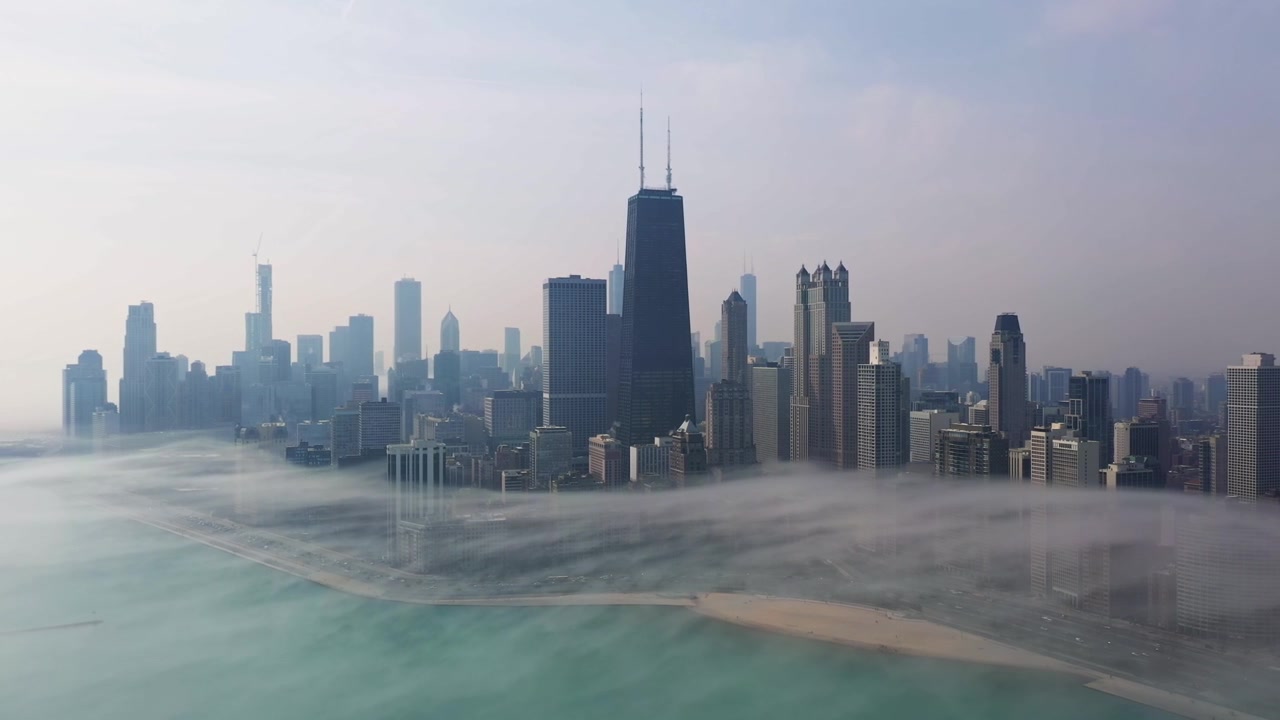 Imagens aéreas suaves movem-se para a esquerda sobre Chicago enquanto a névoa matinal baixa flutua entre a orla do lago e os arranha-céus do centro, criando uma vista atmosférica da cidade.