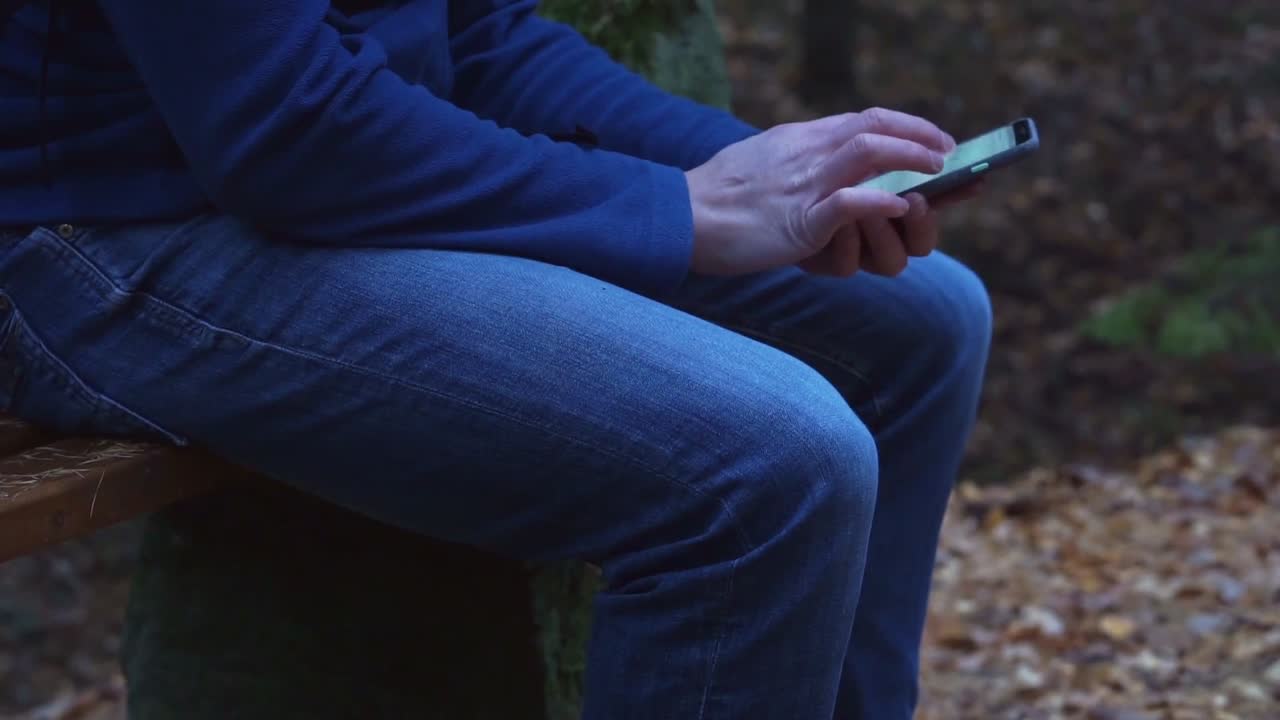 Man in jeans sitting on a bench in forest typing on a smartphone, using apps, closeup of hands, knees and phone