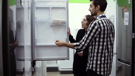 Junge-Ehepaare-Prüfen-Kühlschrank-Mit-Offener-Tür,-Design-Und-Qualität-Vor-Dem-Kauf-In-Einem-Unterhaltungselektronikgeschäft