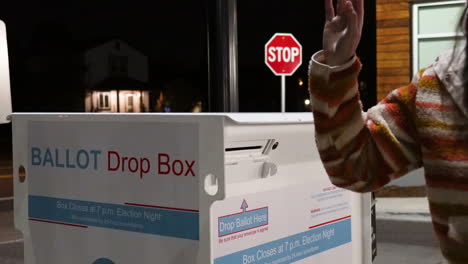 young woman wearing mask drops mail-in ballot letter in slot at official ballot drop box with drop ballot here sign at night