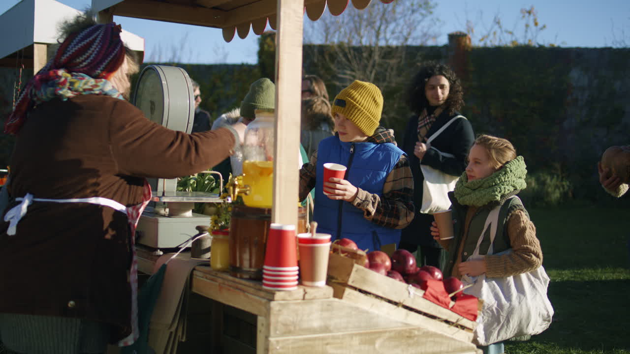 Elderly Woman Pours Drink for Children Elderly Woman at the Stall Pours Lemonade or Tea into Paper Cups for Boy and Girl People Relaxing on Fresh Air on Background Spending Weekend at Local Farmers Market or Autumn Fair Outdoors