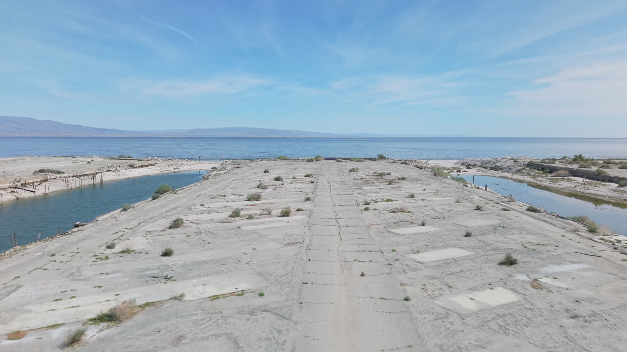 Toma baja de dron volando hacia adelante sobre la marina abandonada en Desert Shores, California, mostrando agua estancada aislada del Mar de Salton a medida que la costa retrocede.