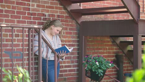 Studentin-Lächelt-Beim-Lesen-Eines-Buches-Draußen-Gegen-Eine-Ziegelwand