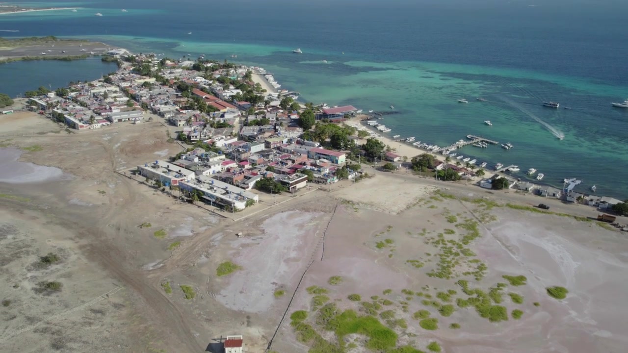 Vista aerea con drone, da zenitale a inclinata verso l'alto, che rivela una vasca di ossidazione, un cimitero locale e la costa di un villaggio di pescatori tropicale sull'isola di Gran Roque, arcipelago caraibico del Venezuela