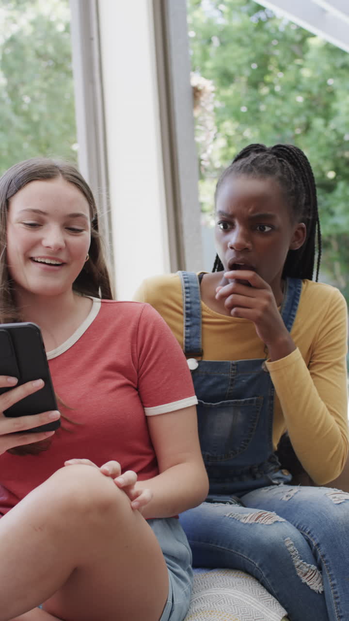 Vertical video of hhappy diverse teenage female friends using smartphone in slow motion