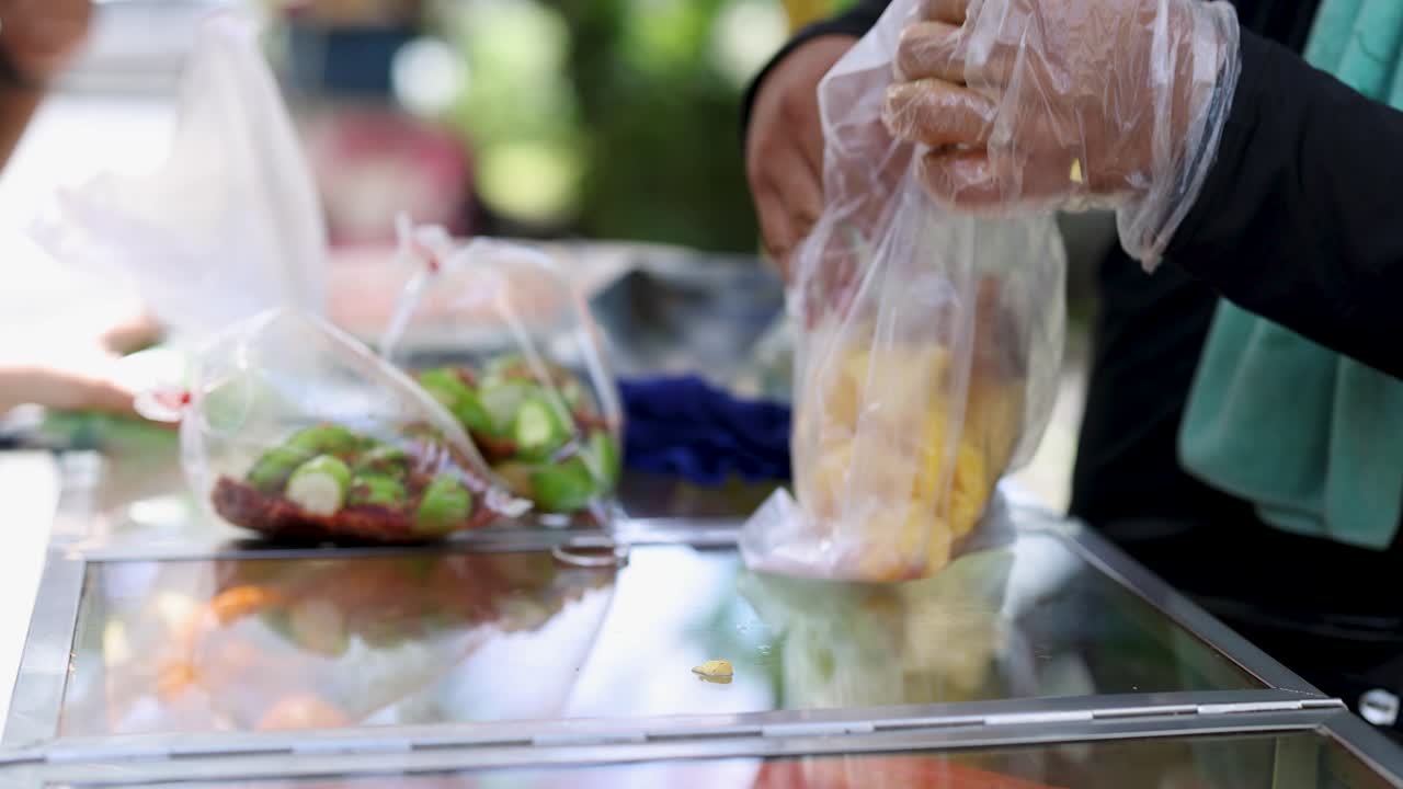 Gloved vendor prepares pineapple with chili sugar in plastic bag at outdoor street food cart