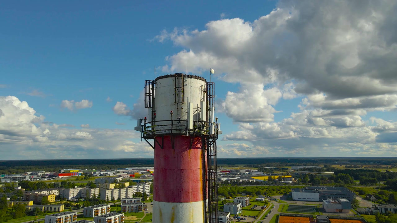 Aerial drone footage orbiting and flying around a tall factory pipe or smoke stack chimney tower that has electronic 5G and 6G radio transmitters on it during a sunny day with urban neighbourhood seen
