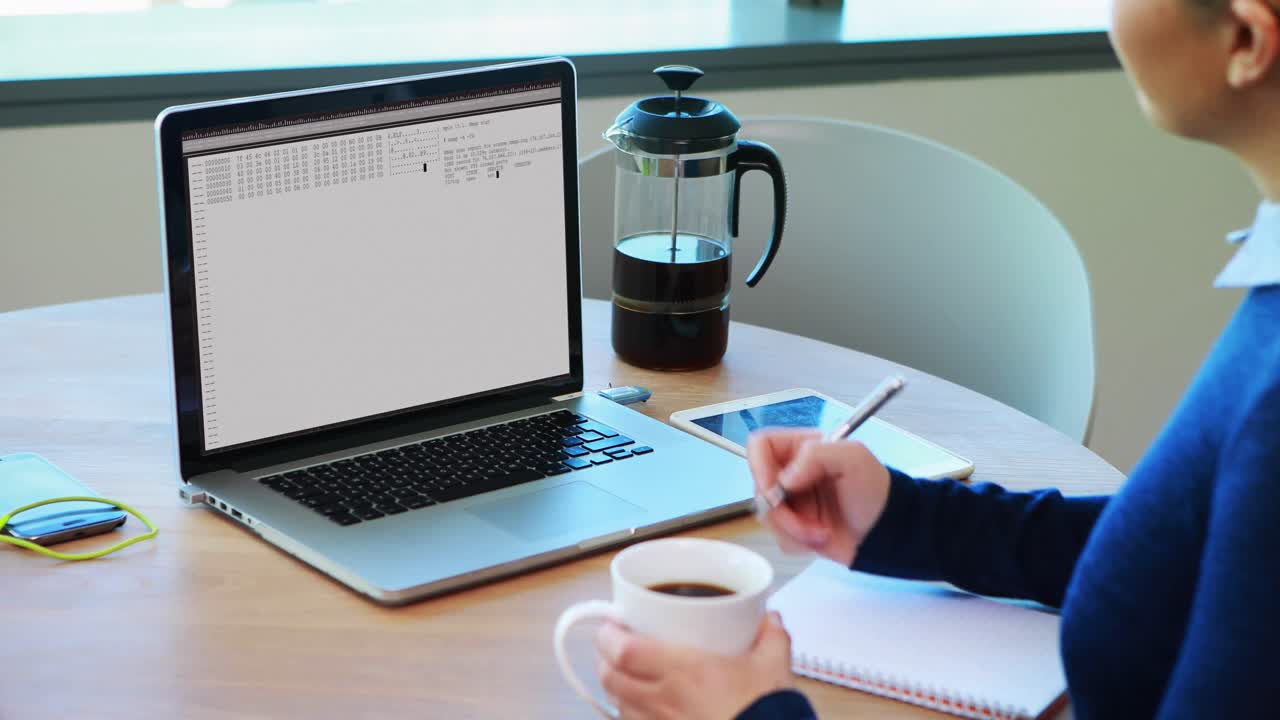 Caucasian woman sitting at table watching coding data processing on laptop screen and making notes