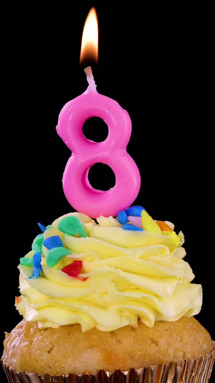 Candela di compleanno rosa numero otto accesa su un cupcake decorato con glassa gialla e codette colorate, su sfondo nero con illuminazione stabile e senza movimento della telecamera.