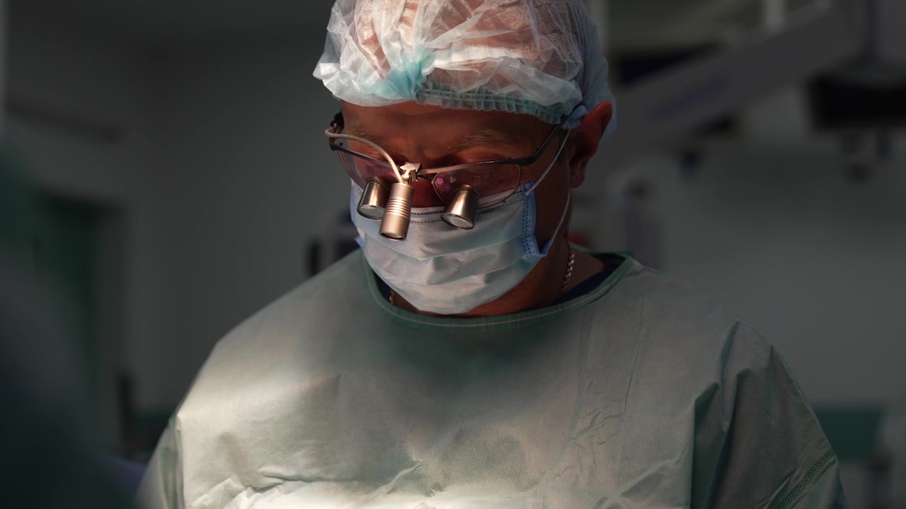 Mandlig kirurg med kirurgiske lupbriller kigger ned på den opererede patient. Portræt af en neurokirurg, der er travlt optaget under operationen.