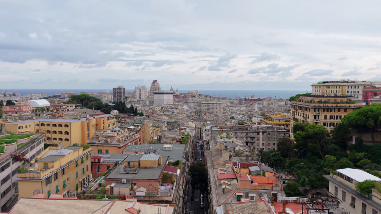 Drone flies forward above the colorful rooftops of Genoa, Italy, showing the historical center, dense urban landscape, cloudy sky, and the sea visible in the distance