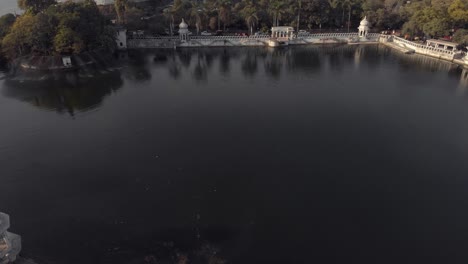 Lake-side-View-from-Gangaur-Ghat-to-The-Leela-Palace-Udaipur-over-Lake-Pichola-in-Udaipur,-Rajasthan,-India---Aerial-Tilt-up-Reveal-shot