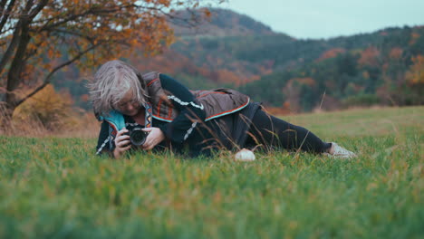 Ältere-Dicke-Frau-Liegt-Mit-Ihrer-Kamera-Im-Gras-In-Der-Natur-Und-Fotografiert-Die-Landschaft,-Umgeben-Von-Bunten-Orangefarbenen-Herbstbäumen,-An-Einem-Kalten,-Windigen-Tag