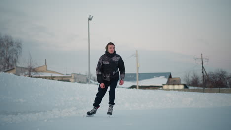 young man skating skillfully on frozen lake near bare trees building and line poles under pale winter sky snow banks frozen surface etched with blade marks reflecting soft light movement energy