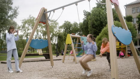 little girl with down syndrome wearing hoodie swinging on a swing in the park on a windy day. her male friend pushing her while her female friend take a photo