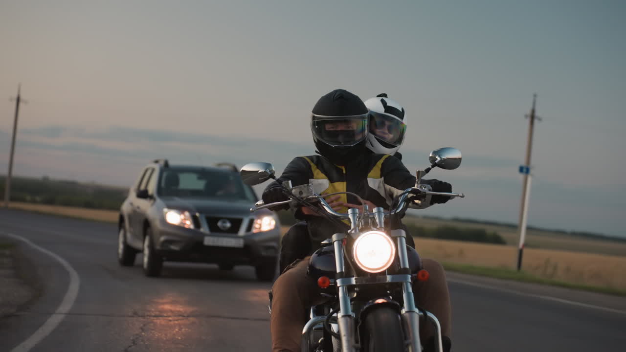 Motorcycle rider with passenger wearing helmets makes bend on rural road while jeep follows close behind, glowing headlight shining against scenic countryside backdrop