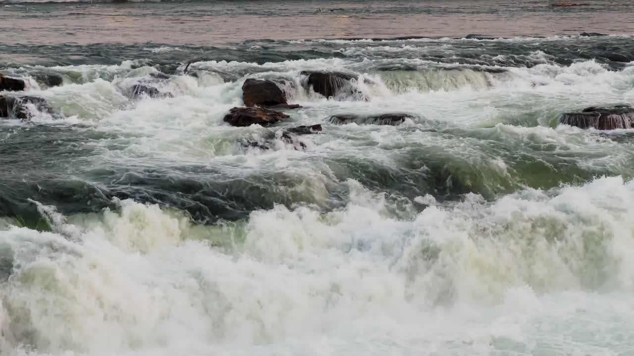 Primo piano di rapide fluviali turbolente che si infrangono sulle rocce, con acqua bianca spumeggiante e forti correnti, catturando il potente movimento naturale all'aperto.