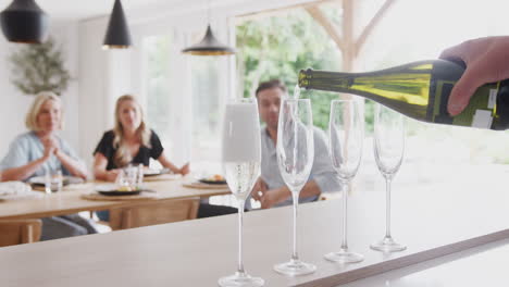 Senior-Man-Opens-Bottle-Of-Champagne-As-Family-With-Adult-Offspring-Eat-Meal-Around-Table-At-Home