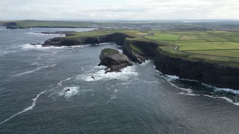 kilkee cliffs in ireland with waves crashing against rocky shores, green fields in the distance, aerial view