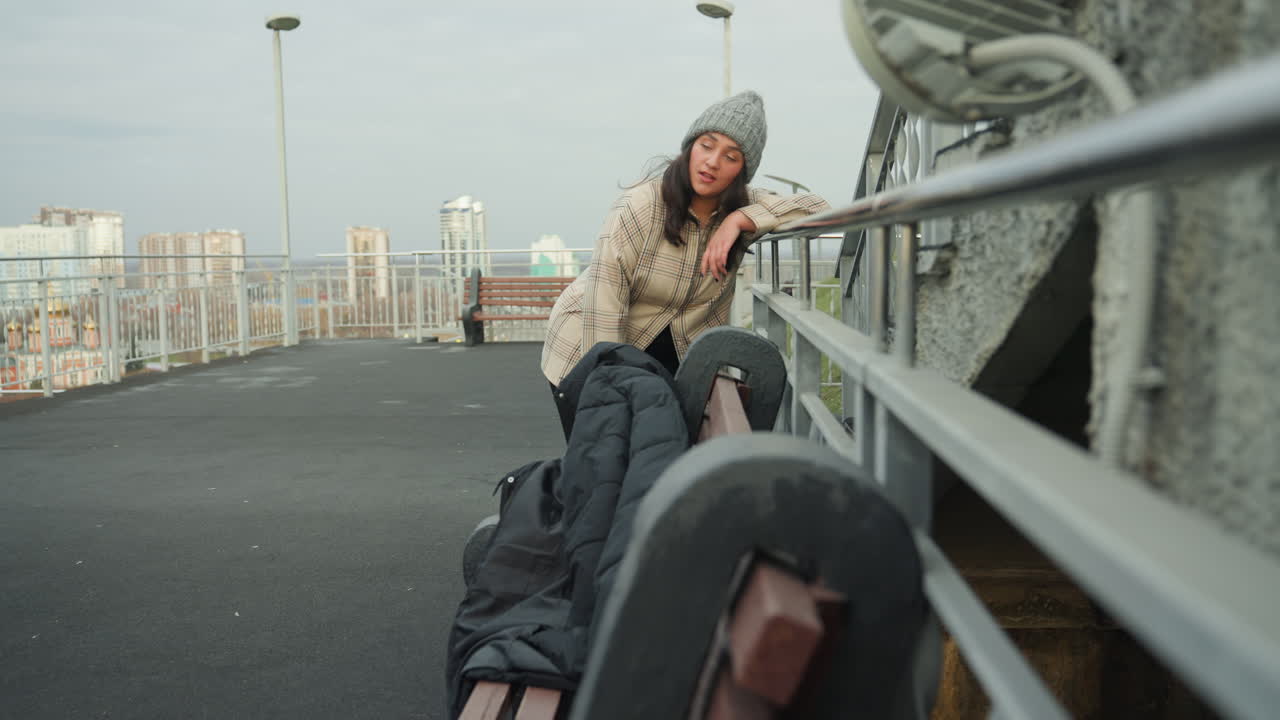 Mujer blanca bailando en una pasarela de azotea con el horizonte de la ciudad, con gorro de punto y abrigo estampado. Mochila y chaqueta en un banco. La escena encuadra su llegada como viajera cruzando un puente y sus pasos expresivos.