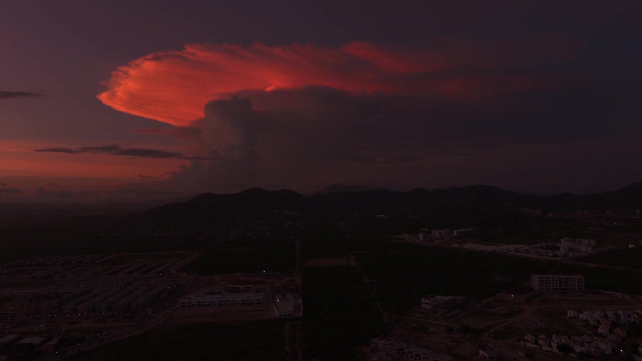 Stunning aerial hyperlapse of a massive colorful storm cloud over the green mountains and cityscape of Cabo San Lucas at dusk.