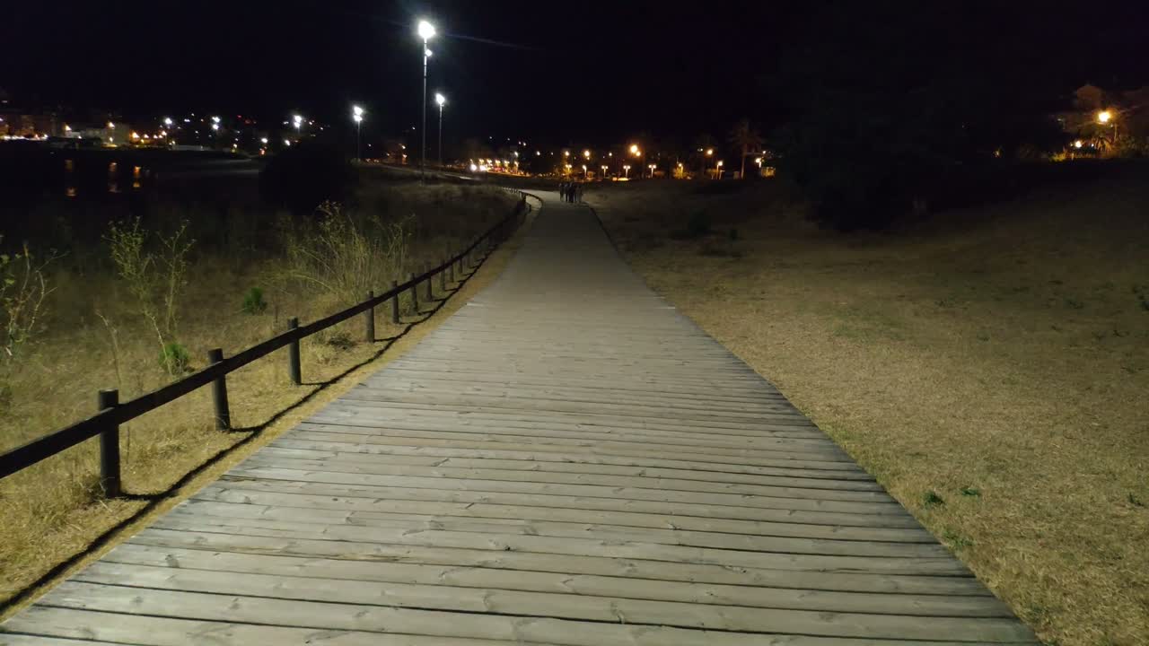 grupo de jóvenes paseando por el paso elevado construido en madera iluminado por farolas durante la noche