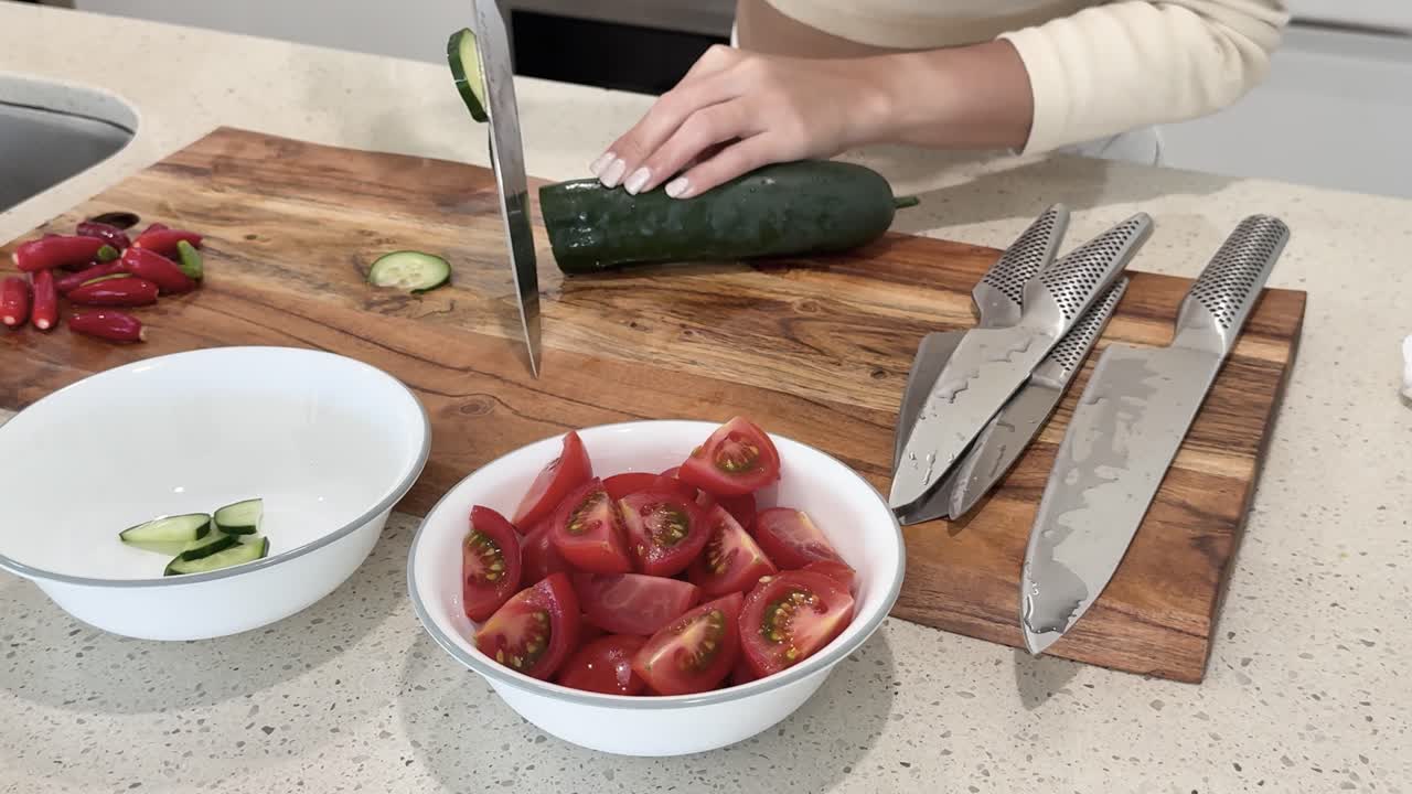 Person slices cucumber on wooden cutting board beside tomatoes in a bright kitchen. Overhead camera angle, natural lighting, clean and organized workspace