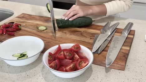 person slices cucumber on wooden cutting board beside tomatoes in a bright kitchen. overhead camera angle, natural lighting, clean and organized workspace