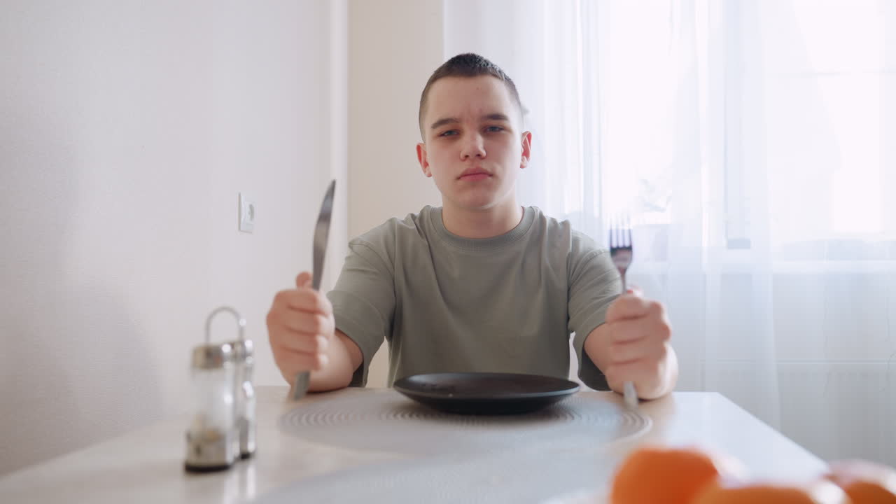 Hungry kid sits at dining table holding spoon and fork, hitting utensils on empty plate with impatient facial expression, waiting for meal in bright home kitchen with sunlight through curtains