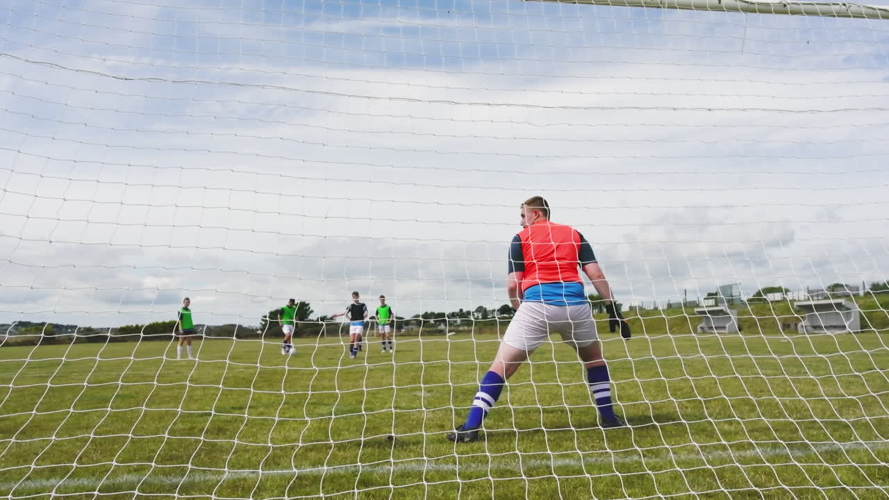 Male and female soccer players playing soccer, shooting at goal with goalkeeper defending on pitch
