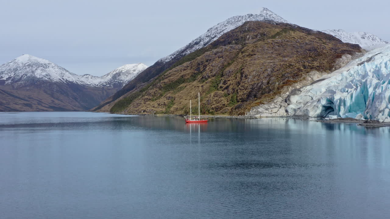 Quiet bay in Patagonia with reflection of hills in calm water under moody overcast sky, aerial pullback from sailboat in turquoise water glaciers