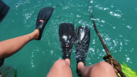 Legs-of-a-couple-sitting-on-the-ledge-of-a-boat-with-scuba-gear-as-they-wait-to-jump-in-the-crystal-clear-blue-water-of-Maracajau-in-Rio-Grande-do-Norte,-Brazil