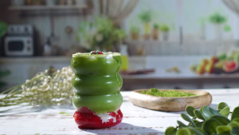 a hand with natural nails gently sets down a vibrant layered matcha strawberry drink in a wavy glass onto a rustic white table, surrounded by green plants and soft natural light.