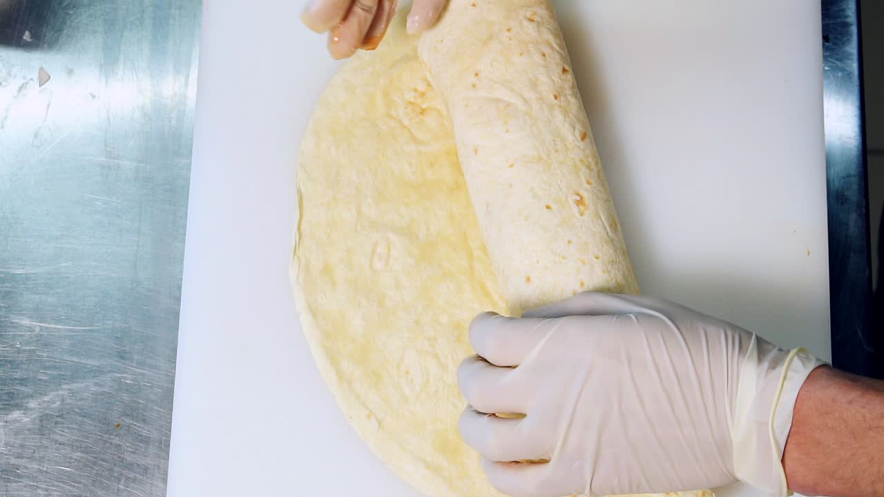 Cook is wrapping pita with filling. Hands of a chef in gloves cooking tasty pita with fresh vegetables. Process of making tasty snack in fast food restaurant. Close-up