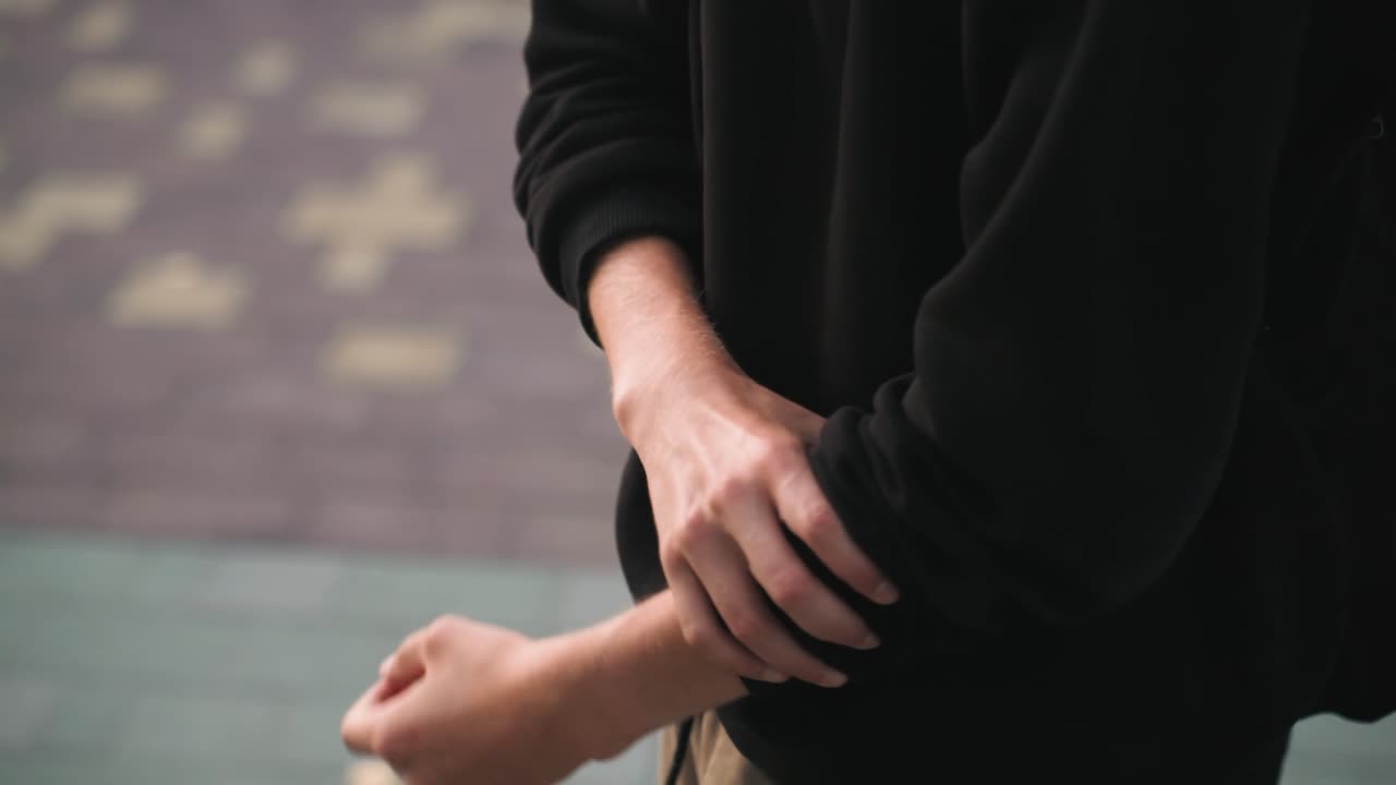 Primo piano di mani caucasiche che sistemano le maniche all'aperto. Uno sfondo di pavimentazione urbana con una maglia casual incornicia le mani che si scaldano, arrotolando i polsini, controllando il polso e stabilizzando il respiro, suggerendo nervosismo.