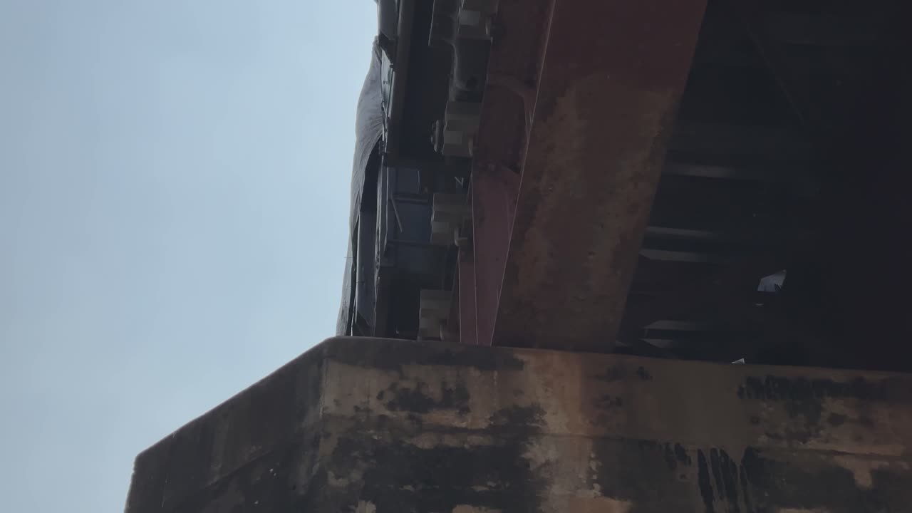 Static shot of a train moving across a rusted metal railway bridge, wheels rattling above the weathered pillar as the structure contrasts against the bright open sky