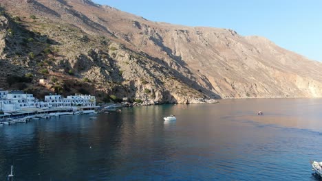 Drone-view-in-Greece-flying-over-blue-sea-in-Loutro-small-white-house-town-and-small-boats-next-to-a-hill-on-a-sunny-day