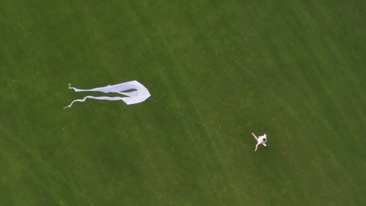 High angle view of a person running across a lush green field while flying a large white kite. Outdoor summer activity and childhood freedom concept