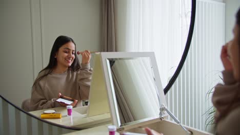 Glückliches-Brünettes-Mädchen-Mit-Grauem-Pullover-Beim-Schminken-Vor-Dem-Spiegel-In-Einem-Modernen-Apartment.-Glückliches-Mädchen-Mit-Brauner-Haut-Macht-Make-up-Und-Trägt-Tagsüber-In-Einem-Hellen,-Modernen-Raum-Puder-Auf-Ihr-Gesicht-Auf